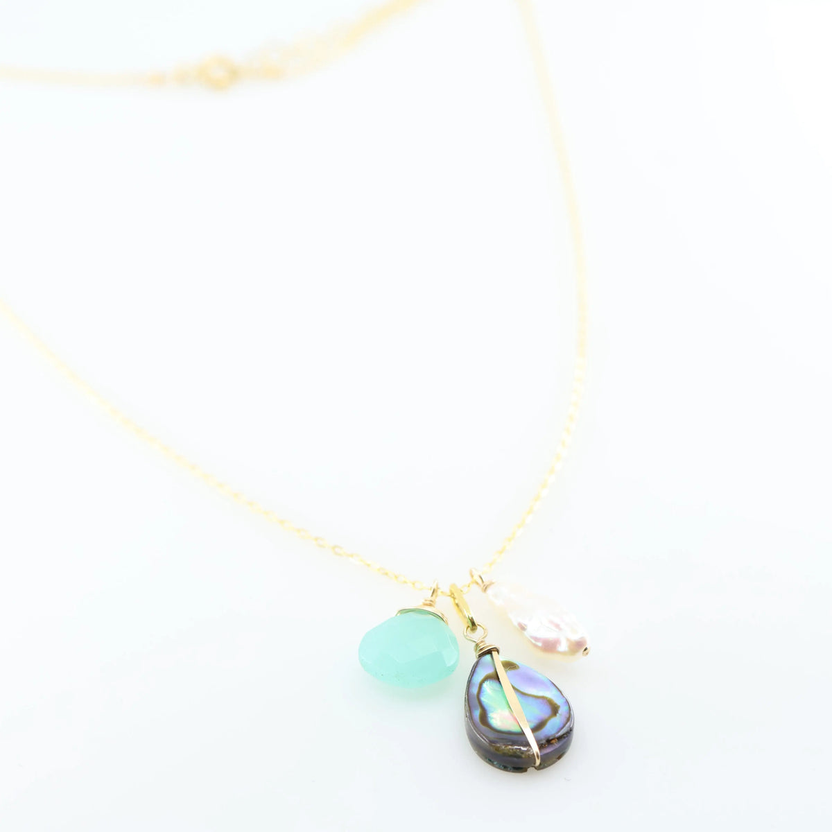 A delicate gold chain necklace featuring three dangling charms: a faceted Dark Aqua Chalcedony gemstone, a teardrop-shaped iridescent abalone shell, and a small Biwa pearl, set against a clean white background.