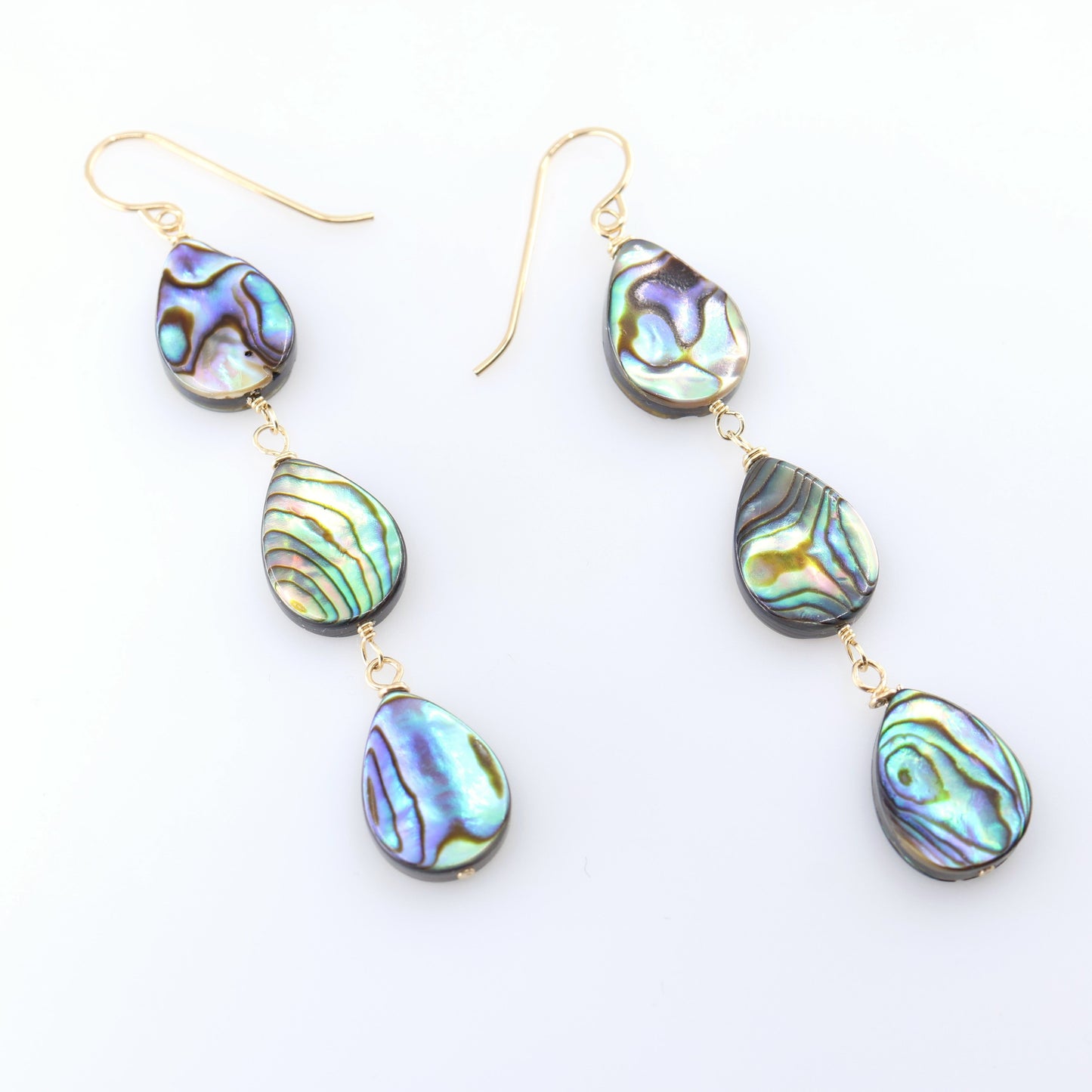 A pair of dangle earrings with three teardrop-shaped abalone shell pendants, showcasing a shimmering iridescent green, blue, and purple coloration, set against a clean white background.