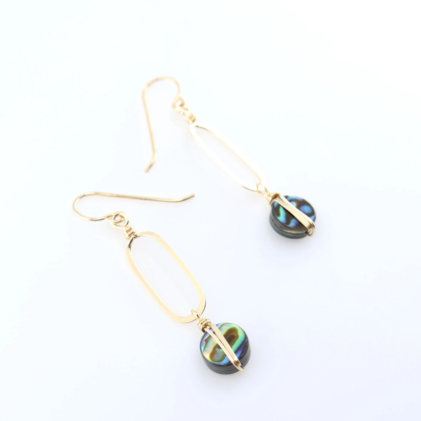 A pair of gold drop earrings with elongated oval hand forged pendant and round iridescent Abalone featuring green, blue, and black swirls, displayed on a clean white background.