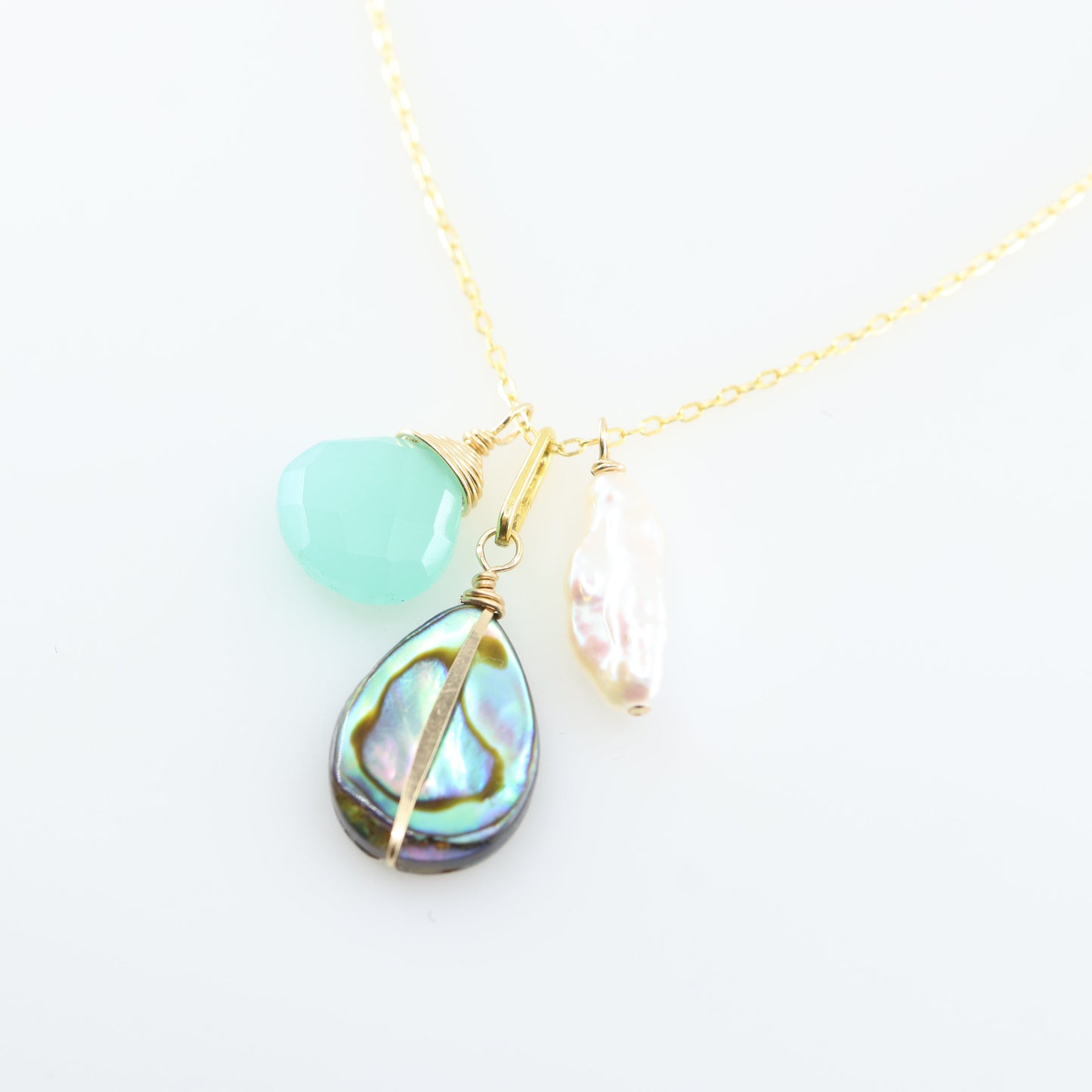 A gold chain necklace featuring three dangling charms: a faceted Dark Aqua Chalcedony gemstone, a teardrop-shaped iridescent abalone shell, and a small Biwa pearl, set against a clean white background.