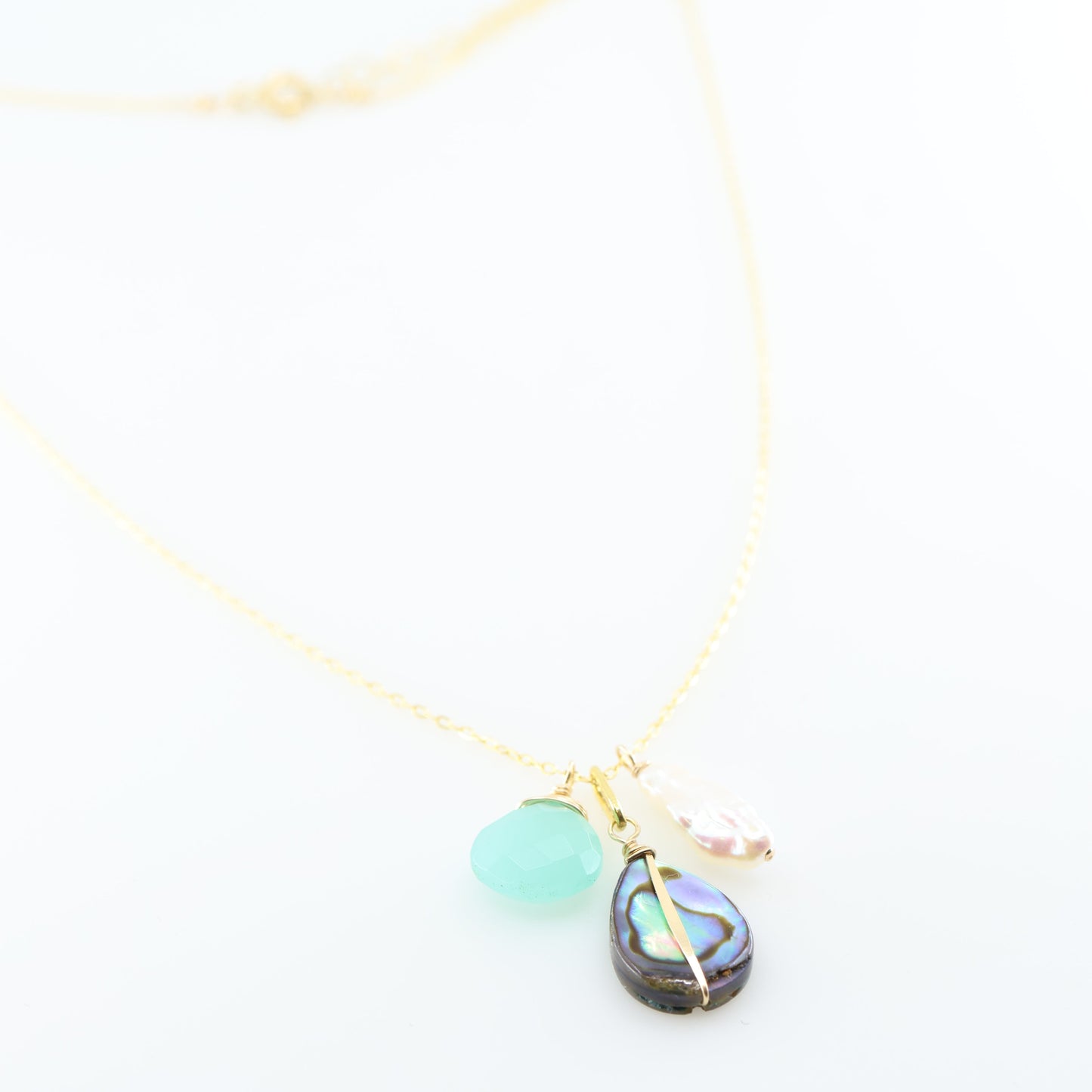 A delicate gold chain necklace featuring three dangling charms: a faceted Dark Aqua Chalcedony gemstone, a teardrop-shaped iridescent abalone shell, and a small Biwa pearl, set against a clean white background.