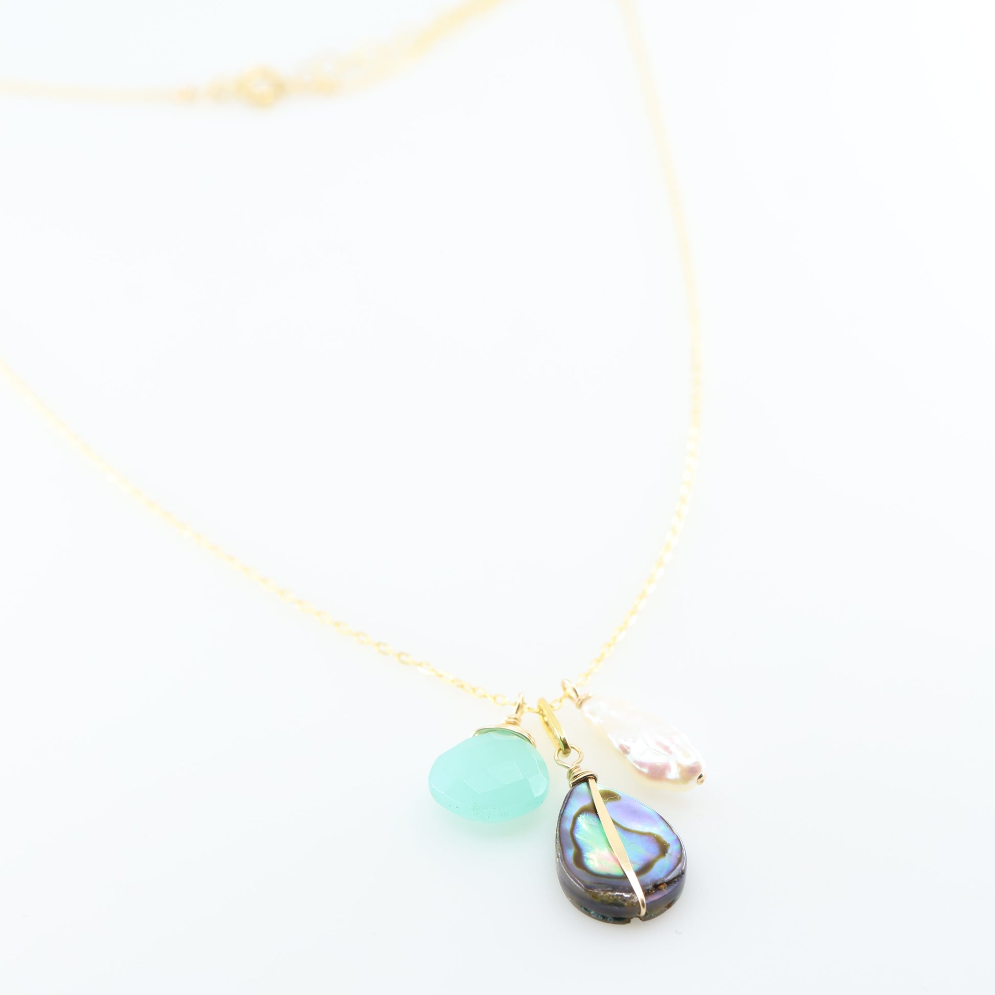 A delicate gold chain necklace featuring three dangling charms: a faceted Dark Aqua Chalcedony gemstone, a teardrop-shaped iridescent abalone shell, and a small Biwa pearl, set against a clean white background.