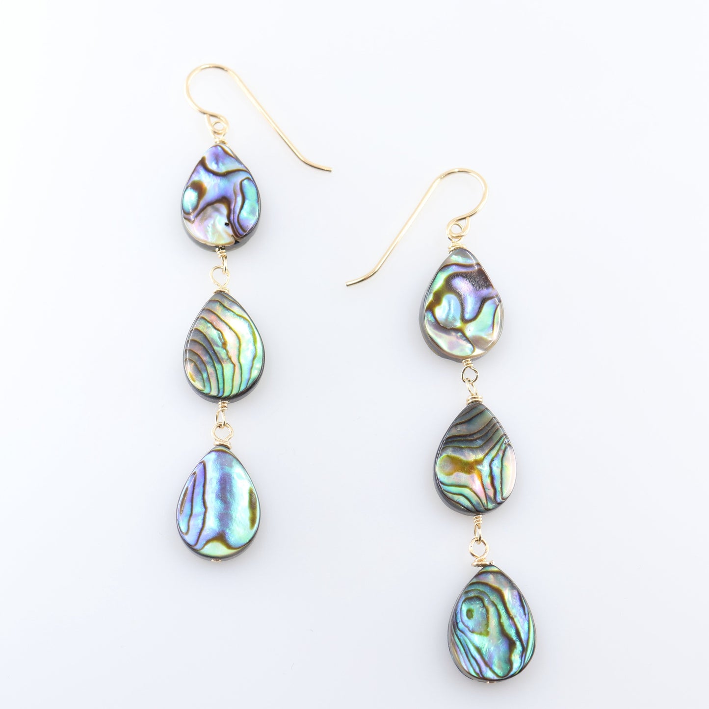 A pair of dangle earrings with three teardrop-shaped abalone shell pendants, showcasing a shimmering iridescent green, blue, and purple coloration, set against a clean white background.