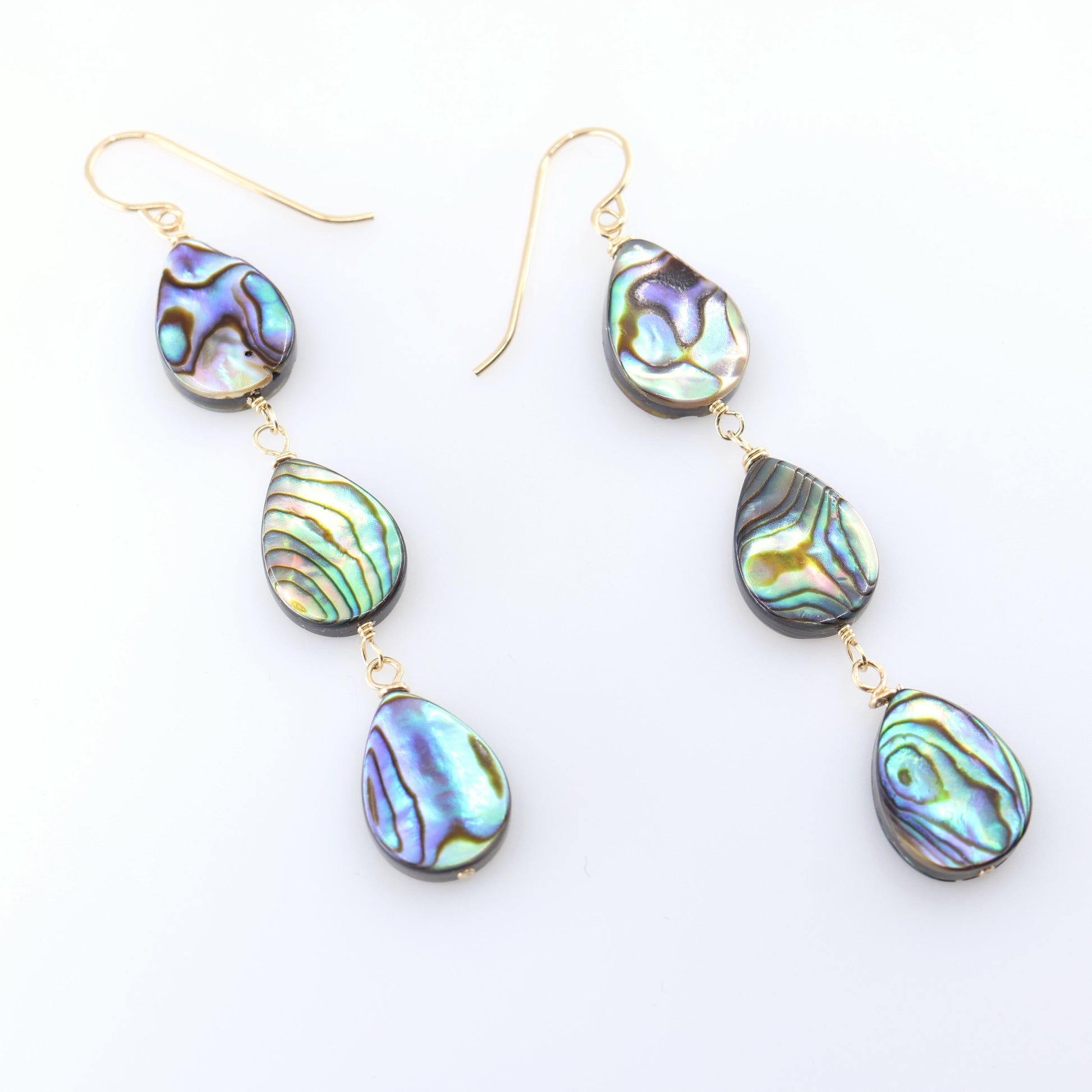 A pair of dangle earrings with three teardrop-shaped abalone shell pendants, showcasing a shimmering iridescent green, blue, and purple coloration, set against a clean white background.