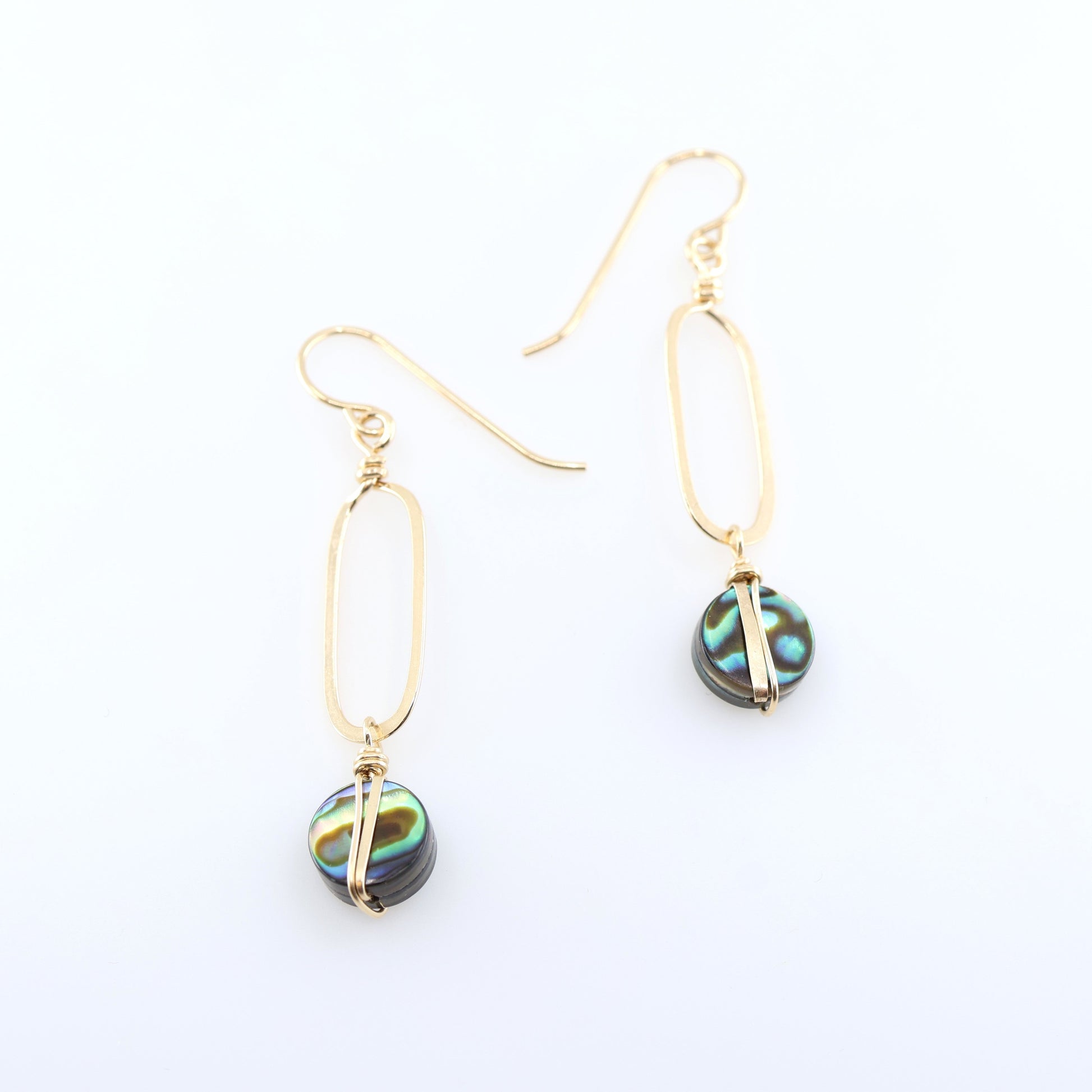 A pair of gold drop earrings with elongated oval hand forged pendant and round iridescent Abalone featuring green, blue, and black swirls, displayed on a clean white background.