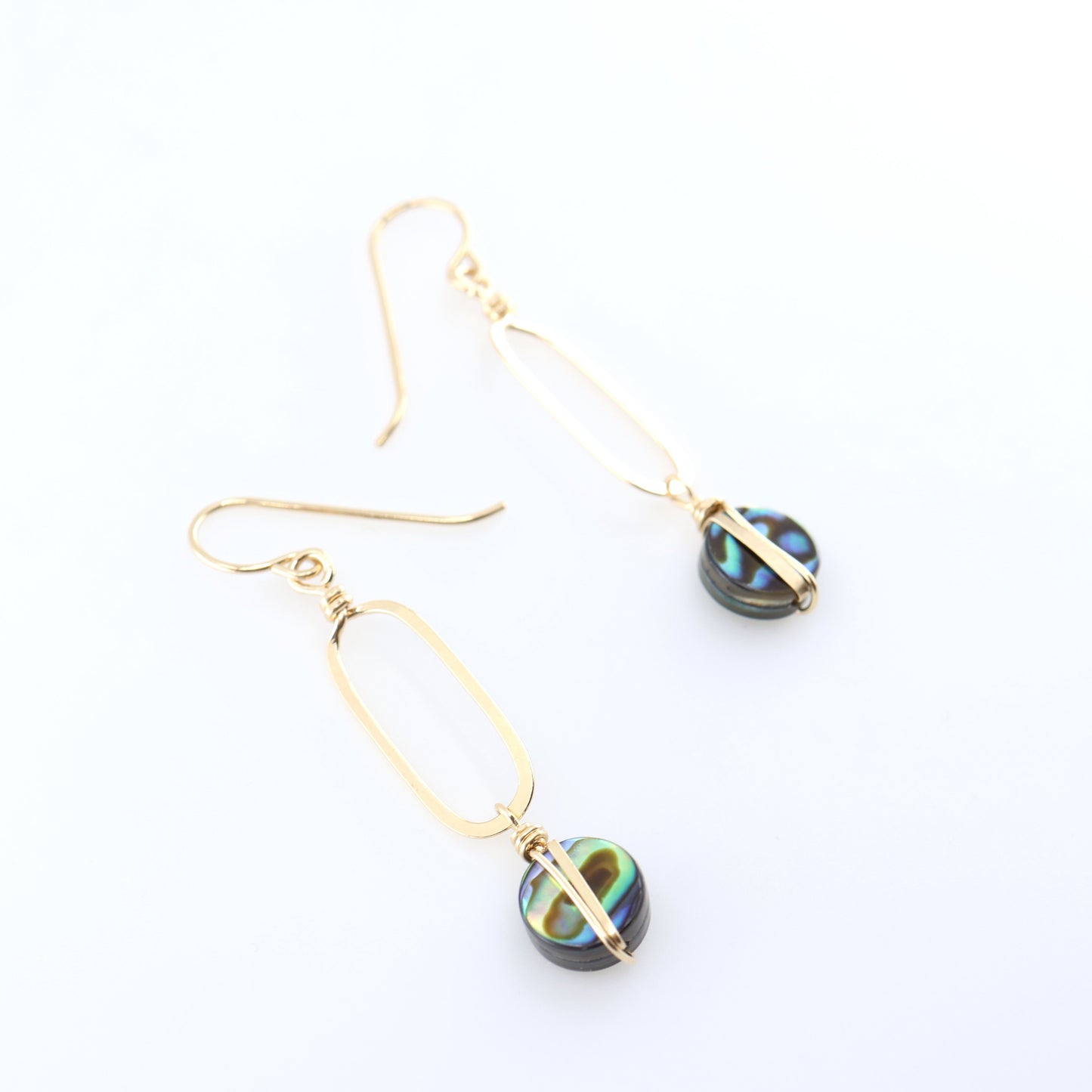 A pair of gold drop earrings with elongated oval hand forged pendant and round iridescent Abalone featuring green, blue, and black swirls, displayed on a clean white background.